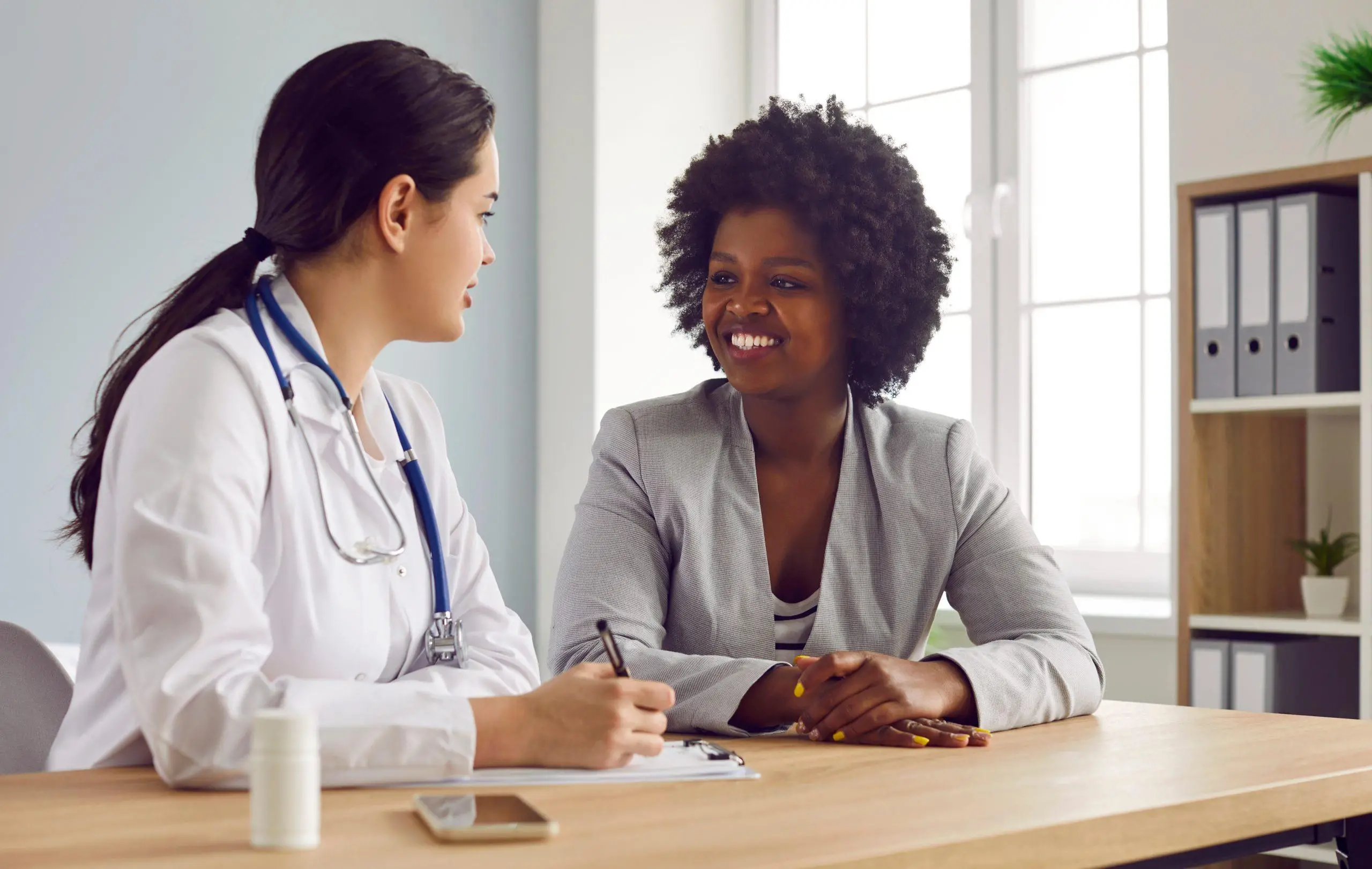 Female friendly doctor talking with a happy smiling african american woman patient and writing down her complaints sitting at the desk in office during medical examination in clinic