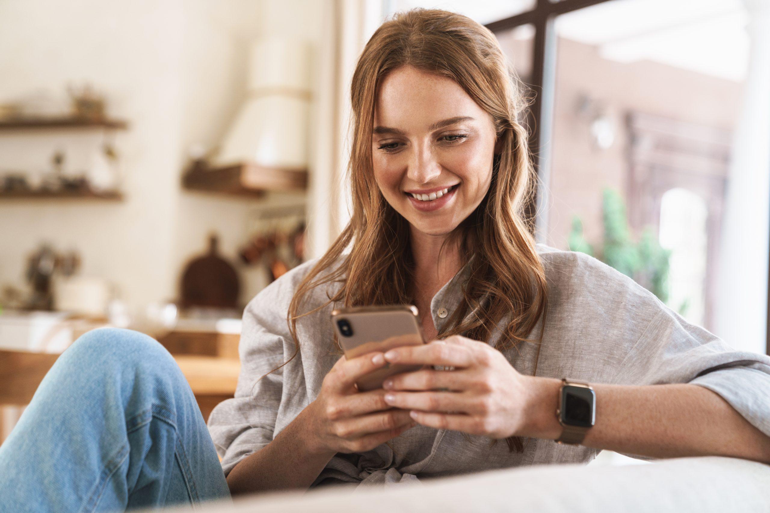 Image of young happy positive smiling cheerful beautiful redhead woman indoors sitting at home using mobile phone.