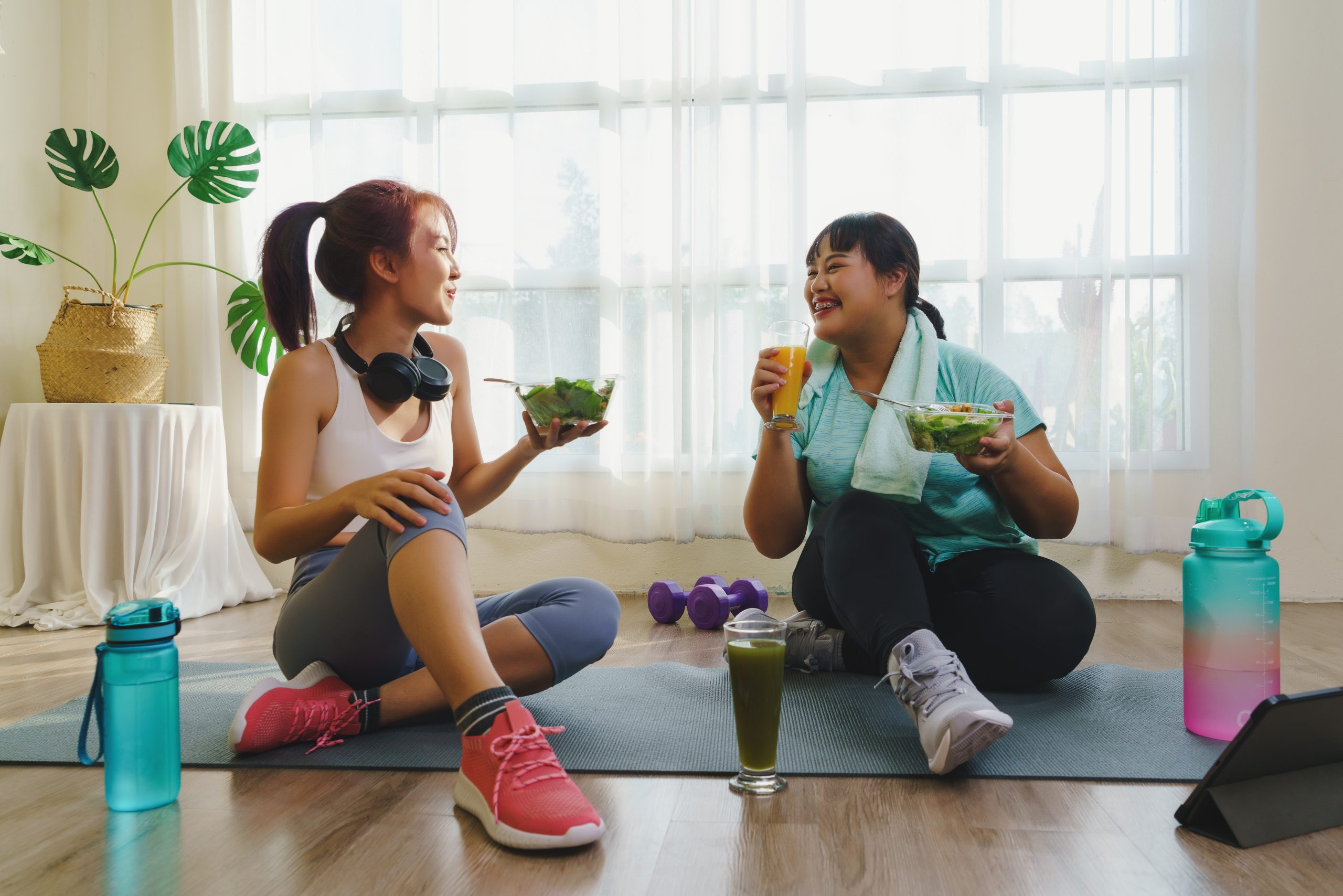 Fitness and Healthy food concept. Two Asian women body size is different in sportswear sitting smiling enjoys while eating healthy food together and happiness after fitness exercising at home.