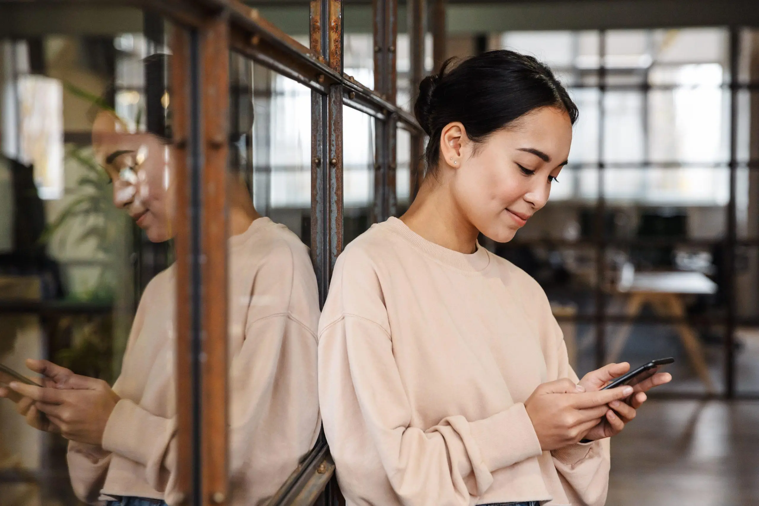 Image of young beautiful asian woman smiling and holding cellphone while working in office