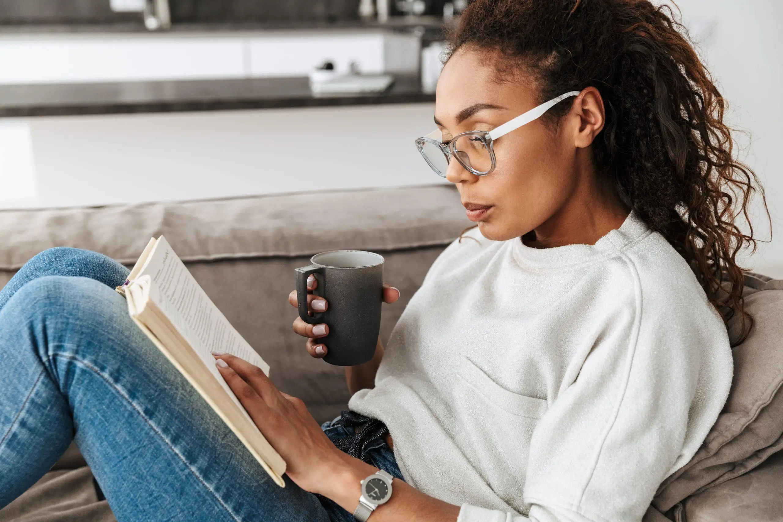 Image of smart african american girl reading book and drinking tea while sitting on sofa in bright flat